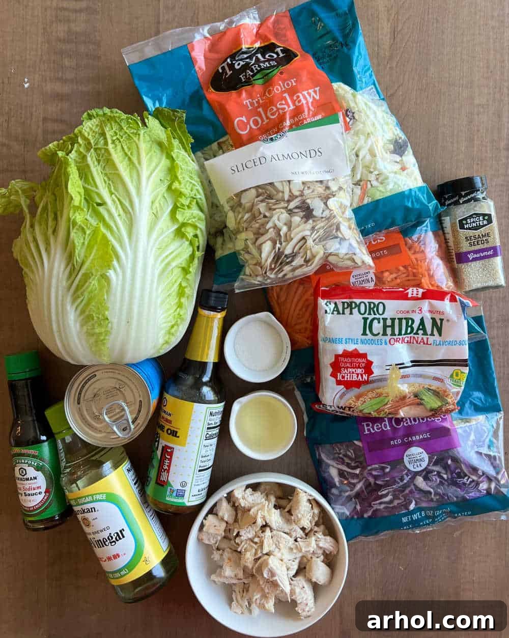 A flat lay image displaying all the fresh ingredients for Chinese Ramen Chicken Salad, including dry ramen noodles, crisp Napa cabbage, colorful coleslaw mix, cooked chicken, blanched almonds, and bright mandarin oranges, alongside dressing components.