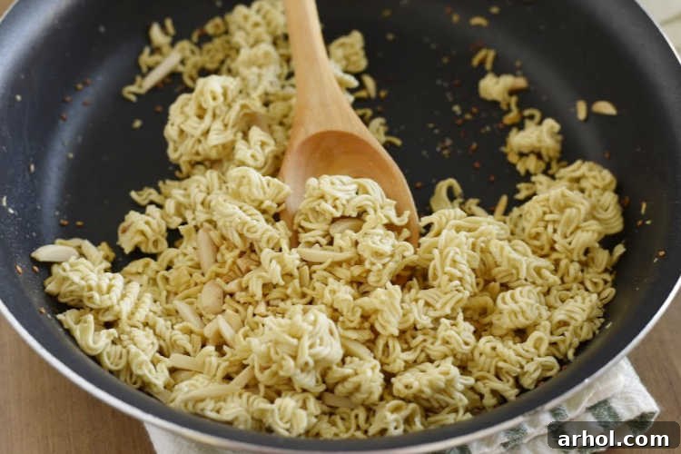 Dry ramen noodles being toasted in a skillet over medium heat, developing a golden brown color and crunchy texture for the salad.