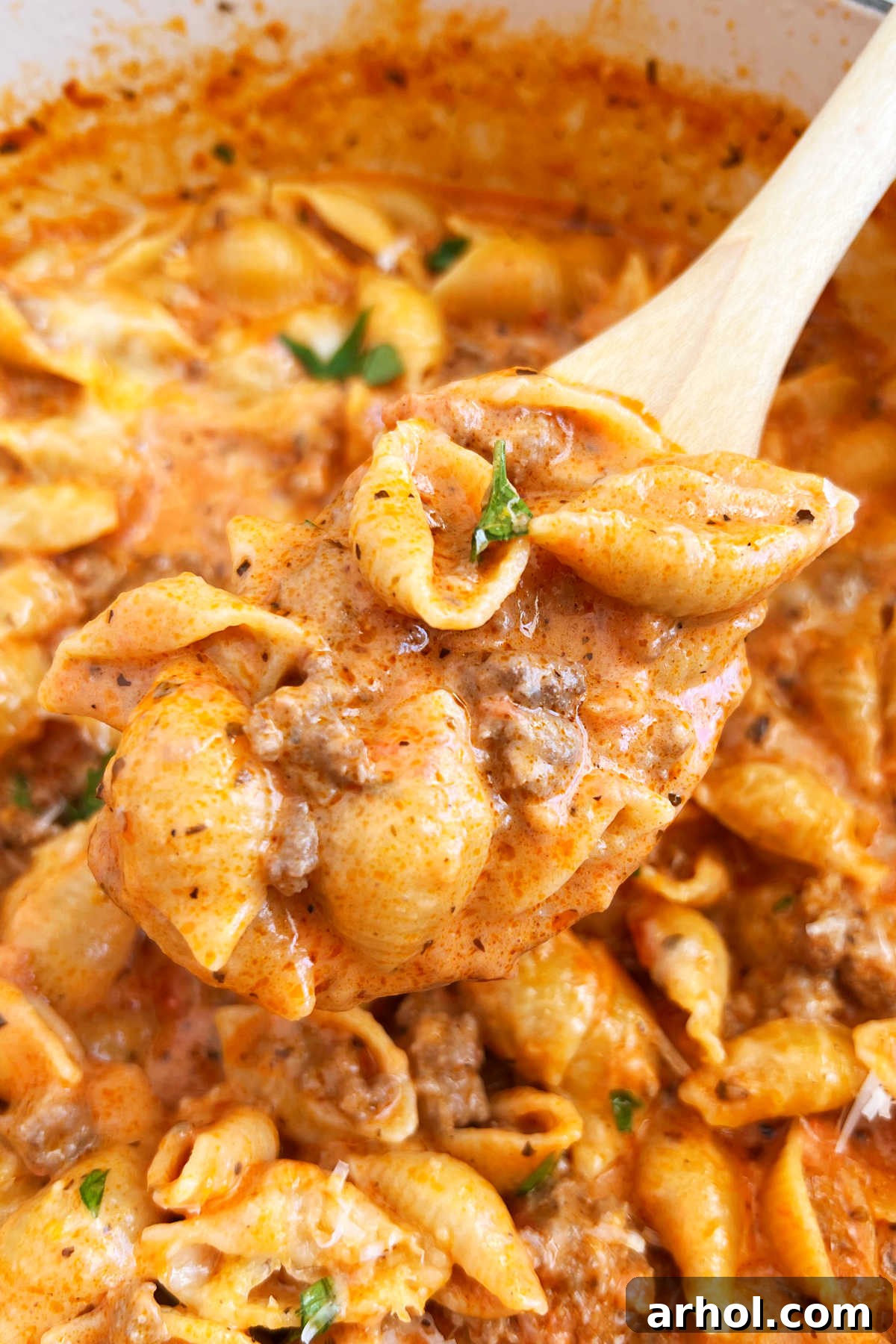 A wooden spoon holding a scoop of creamy shells and beef in tomato sauce.