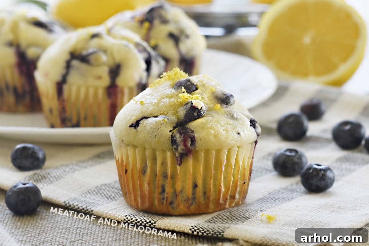 A close-up of a glazed lemon blueberry muffin on a wooden table with fresh blueberries and lemon slices scattered around.