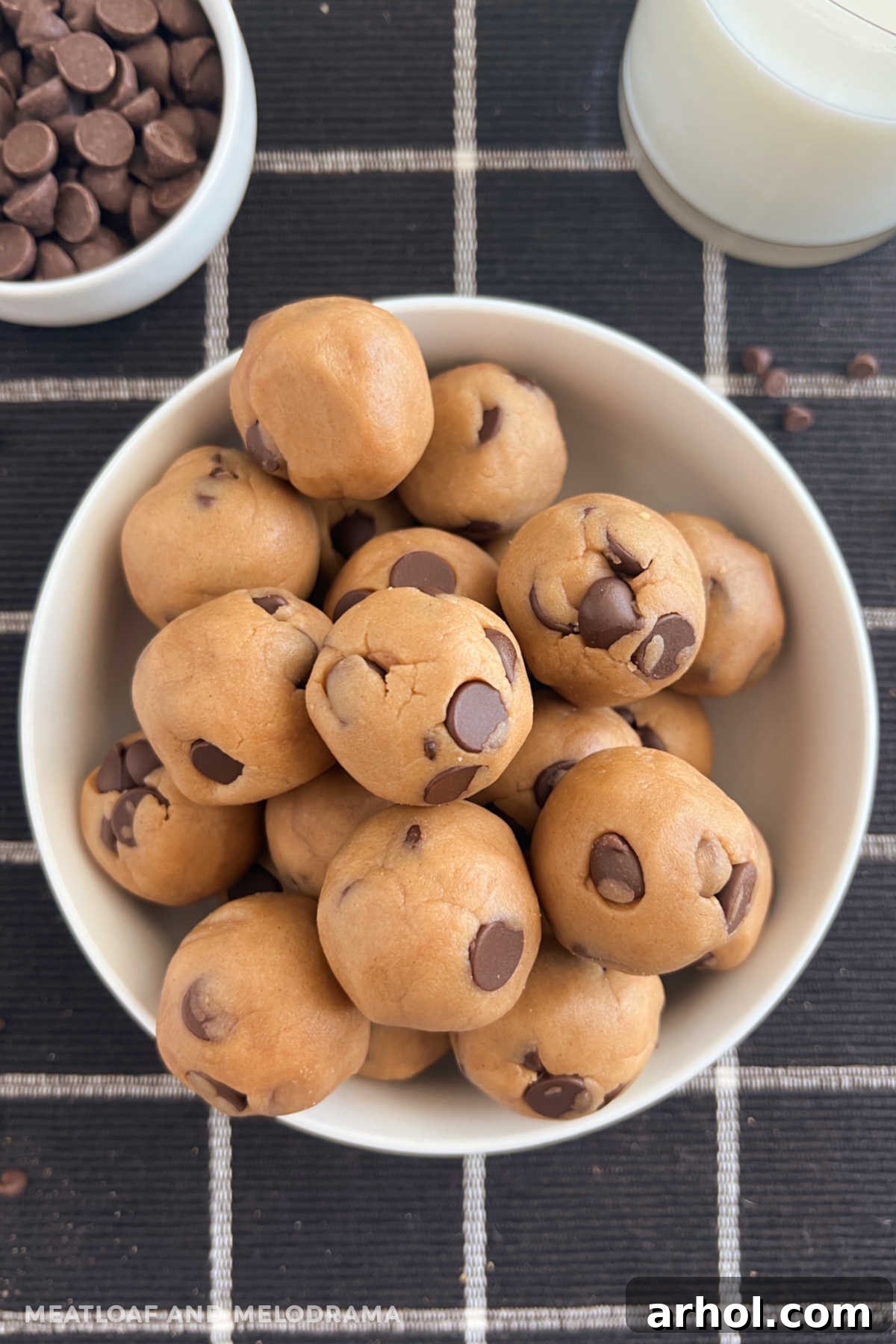 A bowl brimming with no-bake chocolate chip cookie dough balls, accompanied by a glass of milk on a rustic wooden table.