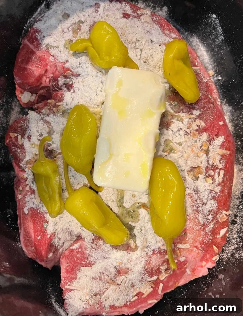 Boneless chuck roast topped with dry ranch dressing, onion soup mix, butter, and peperoncini peppers in a crock pot, ready for cooking