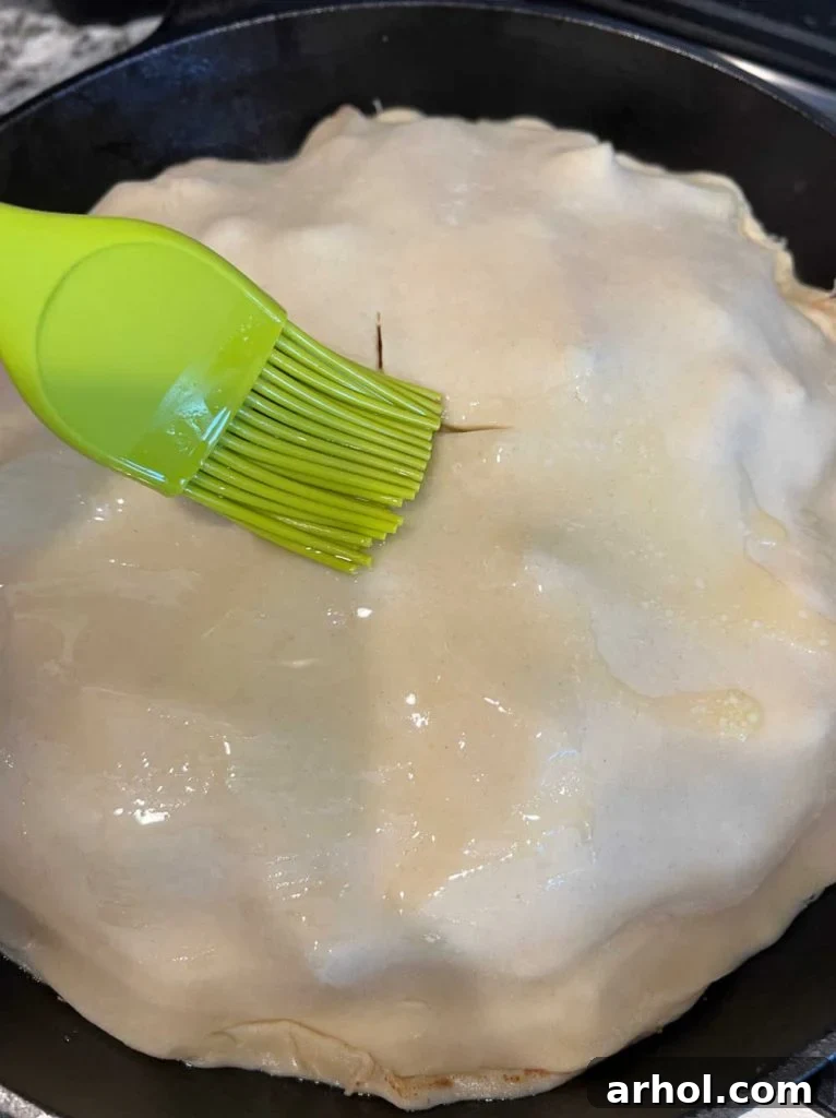 The top pie crust, with steam slits, being brushed with melted butter for a golden finish.