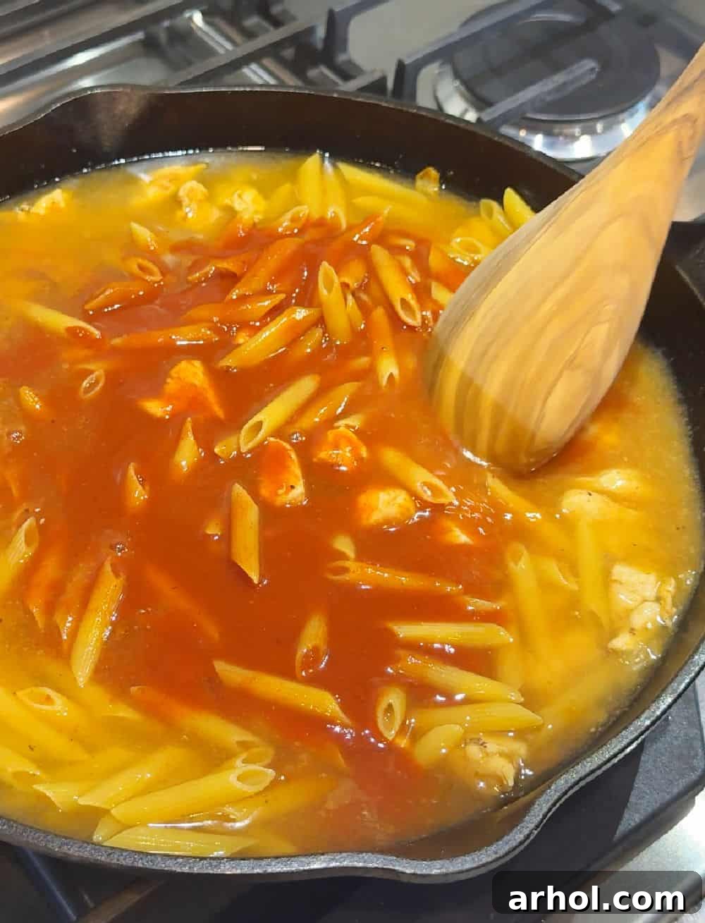 Stirring in water and red enchilada sauce with uncooked pasta and spices in a large skillet, preparing for simmering.