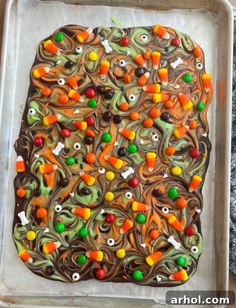 A large sheet of freshly made Halloween candy bark, adorned with various candies and sprinkles, cooling on a baking sheet lined with parchment paper.