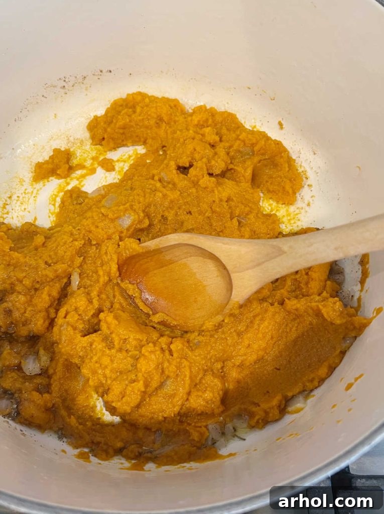 Canned pumpkin puree and spices are being stirred together in a Dutch oven with sautéed onions, gently heating to release flavors.