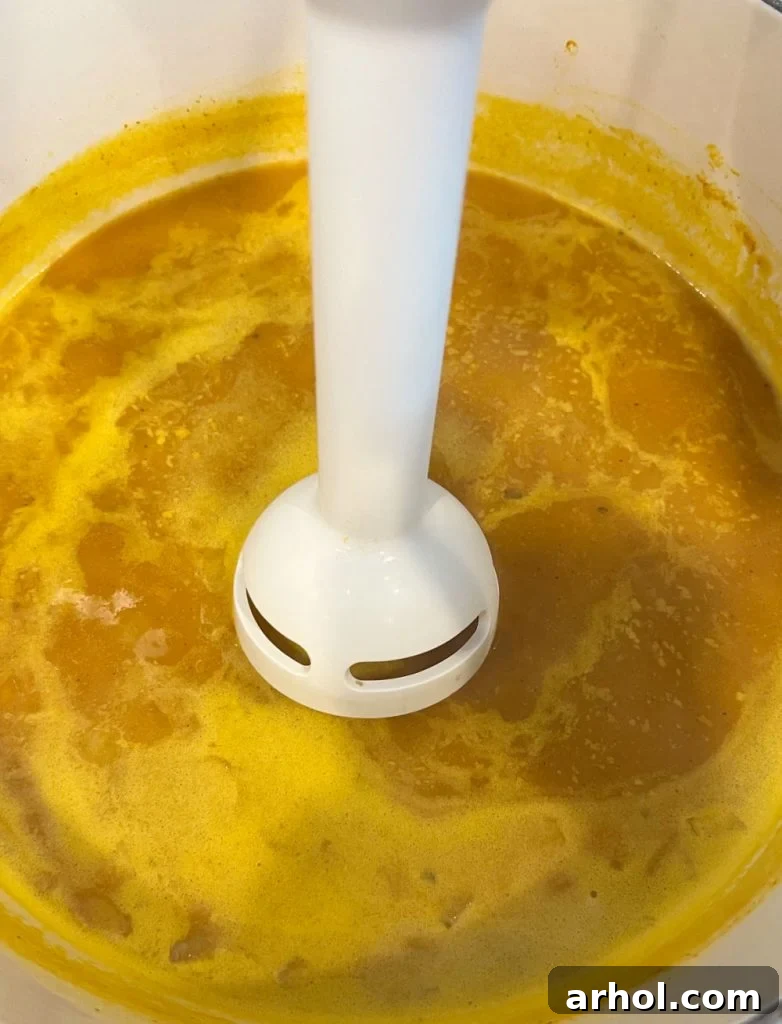 An immersion blender is being used directly in a pot of simmering pumpkin soup, creating a smooth and creamy texture.
