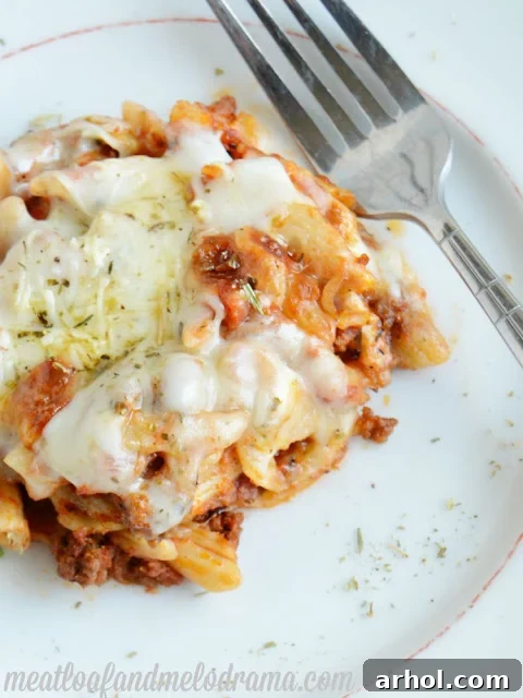 A serving of Cheesy Baked Penne Pasta on a white plate, garnished with fresh parsley, showing the rich texture of the pasta, beef, and melted cheese.