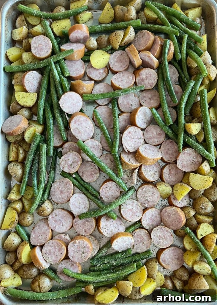 Roasted baby potatoes with fresh green beans and sliced sausage coins arranged on a sheet pan, halfway through cooking.