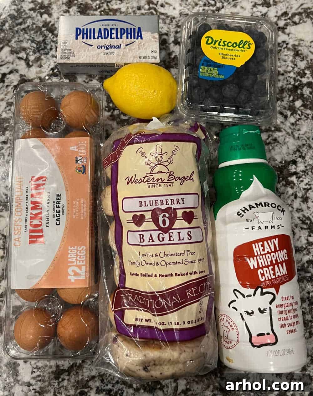 Close-up shot of key ingredients for the Lemon Blueberry Bagel Breakfast Casserole, including blueberry bagels, fresh eggs, cream cheese, a lemon, blueberries, and heavy cream, all neatly arranged.