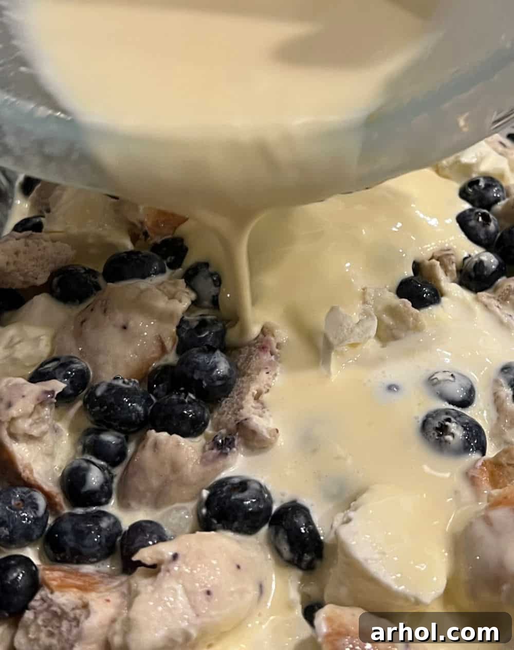 The lemon custard being poured over the bagel and blueberry mixture in a baking dish, preparing for refrigeration.