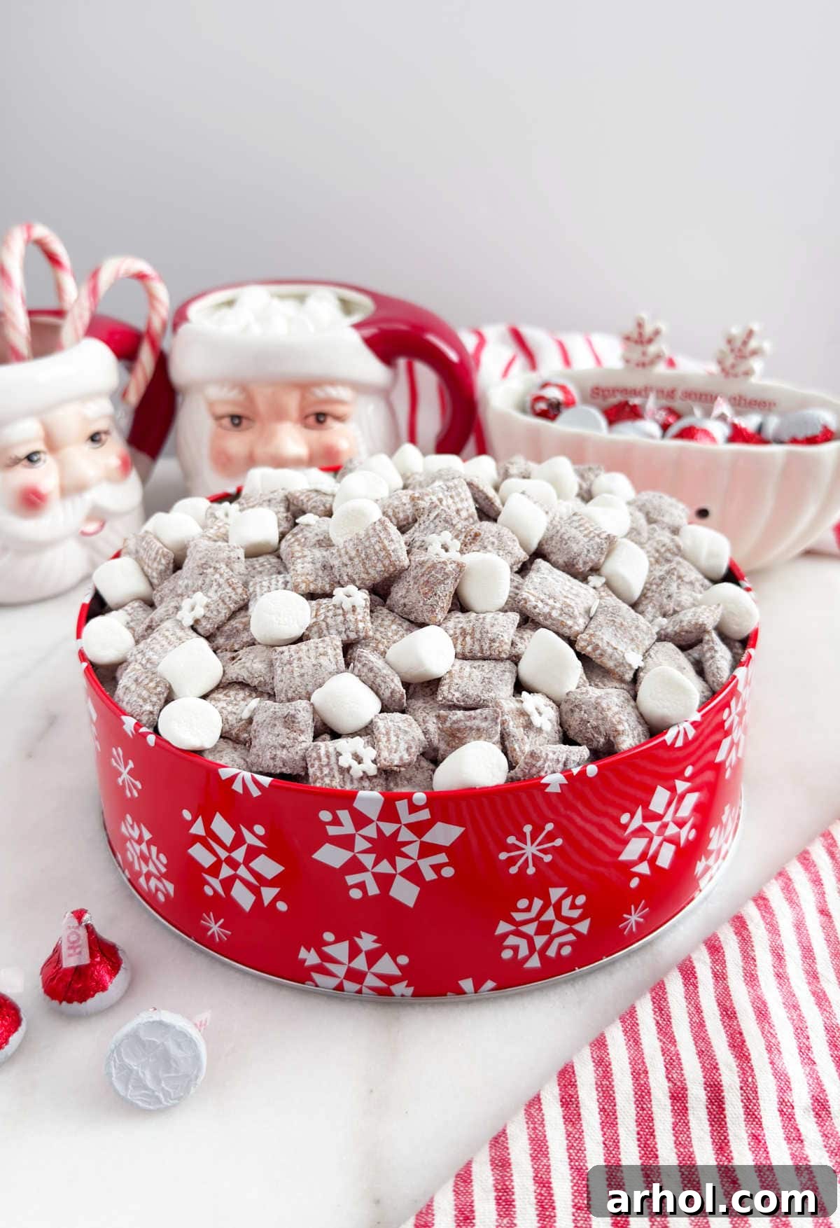 A vibrant red tin overflows with Hot Chocolate Muddy Buddies, also known as puppy chow, garnished with fluffy mini marshmallows.