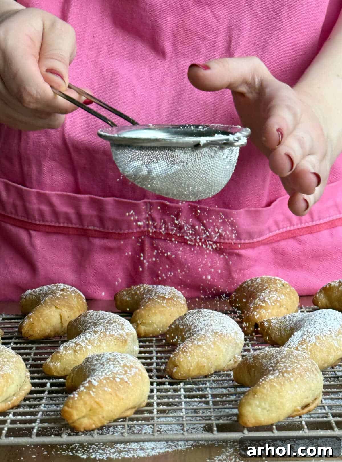 Dusting baked nut horn cookies generously with powdered sugar on a cooling rack.