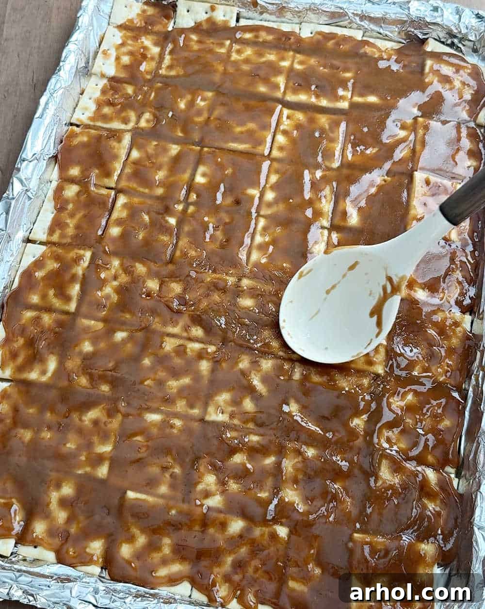 Image depicting the hot toffee mixture being spread evenly over a layer of saltine crackers on a baking sheet.