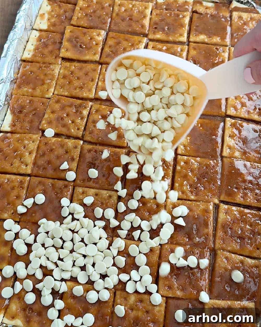 Image illustrating white chocolate chips being sprinkled generously over the hot toffee layer on the crackers.
