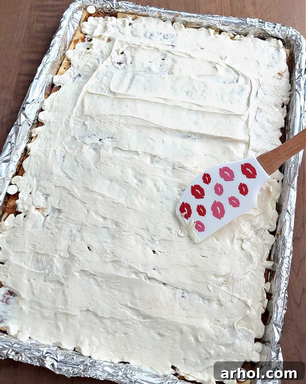 Image demonstrating a spatula smoothing the melted white chocolate evenly across the crackers, covering the toffee.