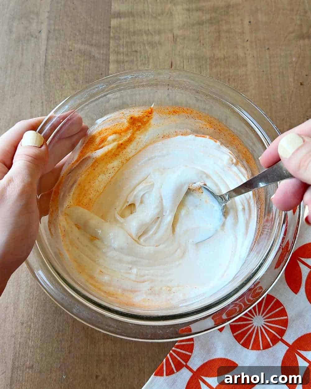 A hand mixing the ghost pepper puree into the mayonnaise and sour cream mixture in a small glass bowl with a whisk, ensuring a smooth blend.