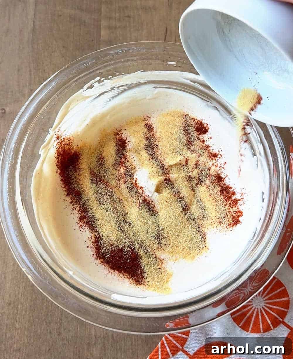 A hand adding a spoonful of powdered spices, including garlic powder and paprika, into the creamy dip mixture, enhancing its flavor profile.