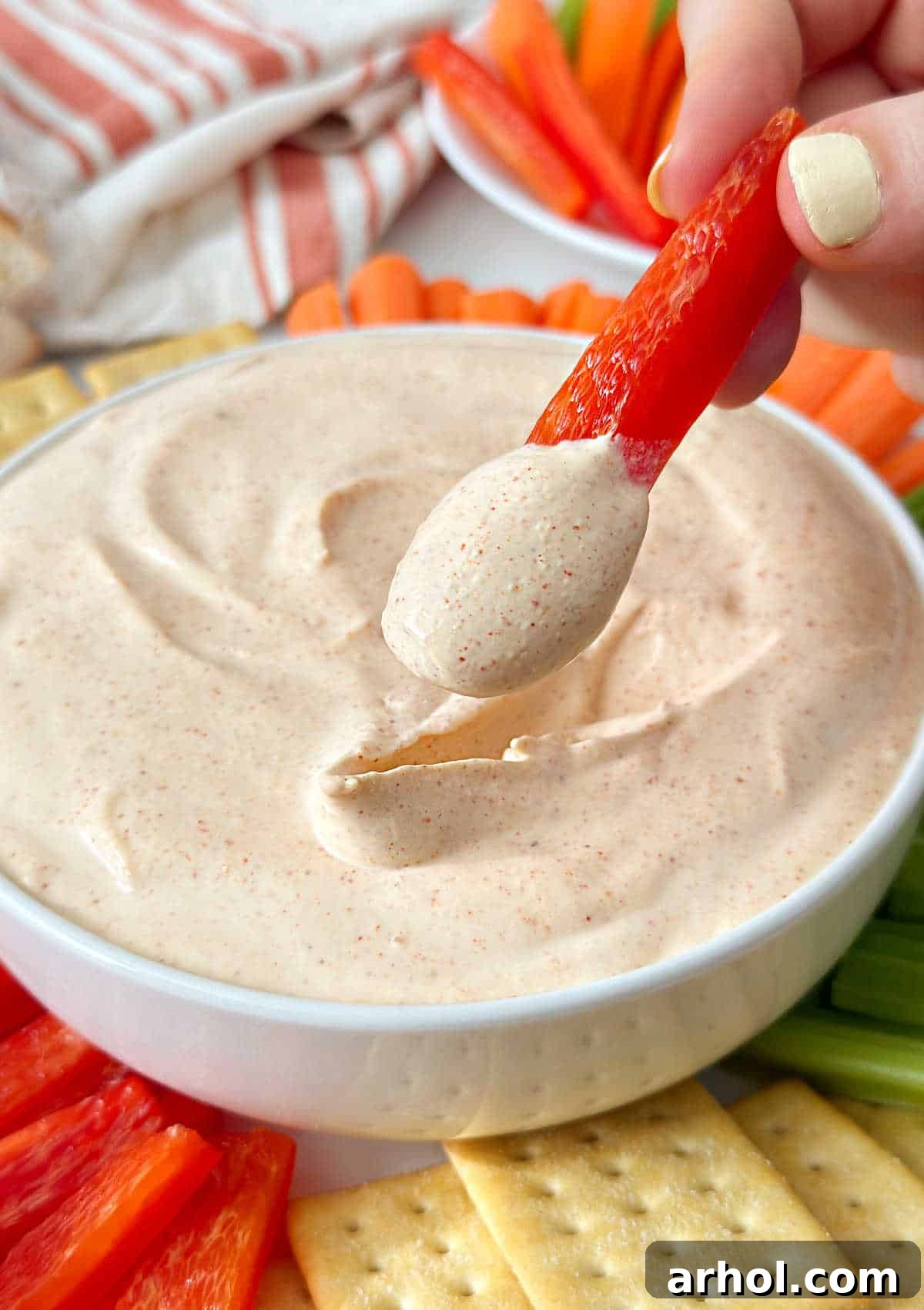 A hand dipping a vibrant slice of red bell pepper into a bowl of creamy ghost pepper dip, ready to enjoy a spicy and refreshing bite.