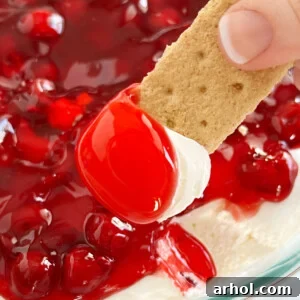 A generous graham cracker dipped into a luscious Cherry Cheesecake Dip, showcasing the sweet pie filling on top, ready to be enjoyed.
