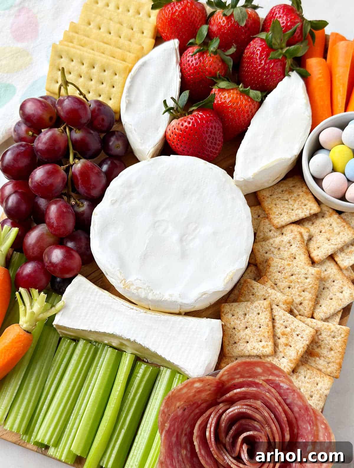 Delightful Easter charcuterie board featuring a brie cheese bunny, a decorative salami rose, an array of fresh vegetables, vibrant fruits, colorful candy eggs, and various crackers arranged for a festive spring gathering.