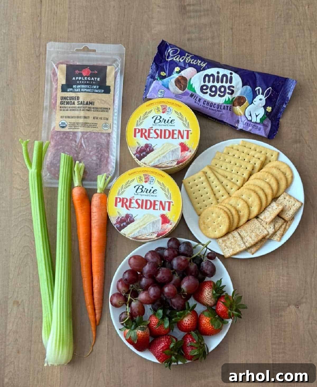 A flat lay image showcasing all the essential ingredients for an Easter charcuterie board: salami slices, two wheels of brie cheese, assorted candy eggs, a variety of crackers, fresh red grapes and strawberries, and crisp carrots and celery sticks.
