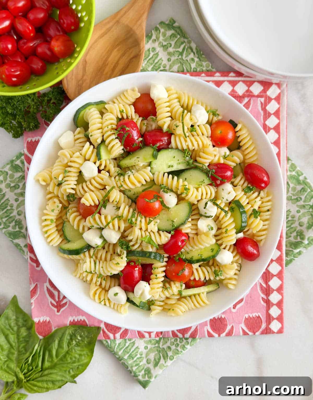Vibrant cucumber tomato pasta salad with fresh basil and mozzarella pearls in a white ceramic bowl, set on a rustic wooden table.