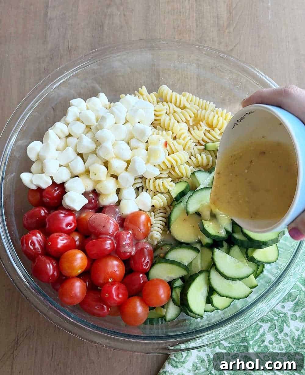 Step 2: Pouring vibrant Italian dressing generously over a bowl filled with pasta, cucumbers, and tomatoes, preparing for a thorough toss.