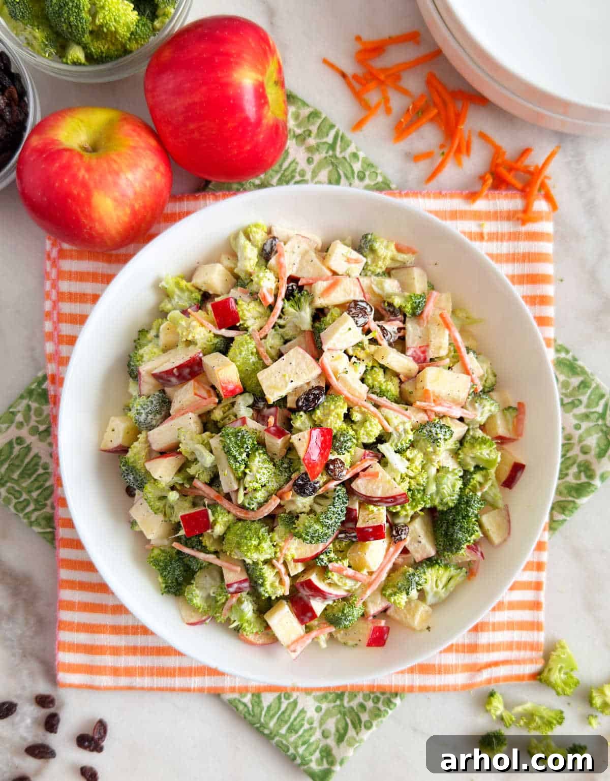 A large white bowl filled with vibrant broccoli apple salad, featuring green broccoli, red apple pieces, orange carrots, and dark raisins, ready to be served on a rustic wooden table.