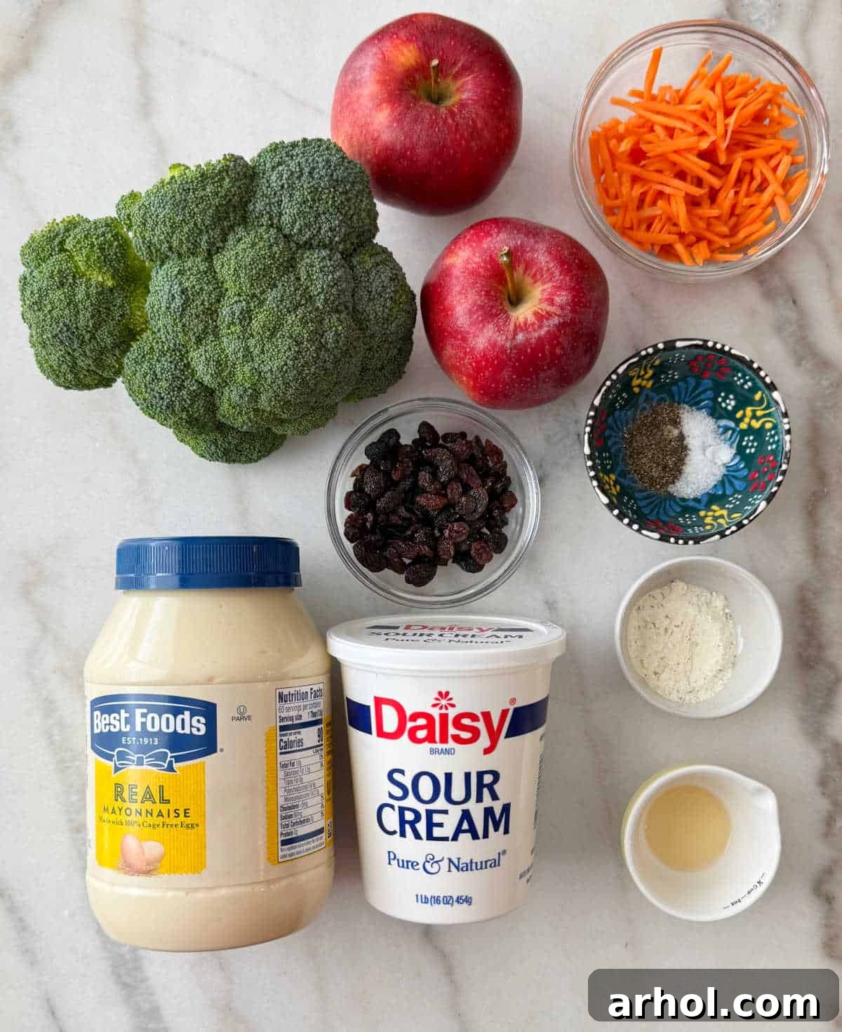 All the ingredients for broccoli apple salad laid out: mayonnaise, sour cream, fresh broccoli, diced apples, shredded carrots, raisins, apple cider vinegar, and ranch seasoning.