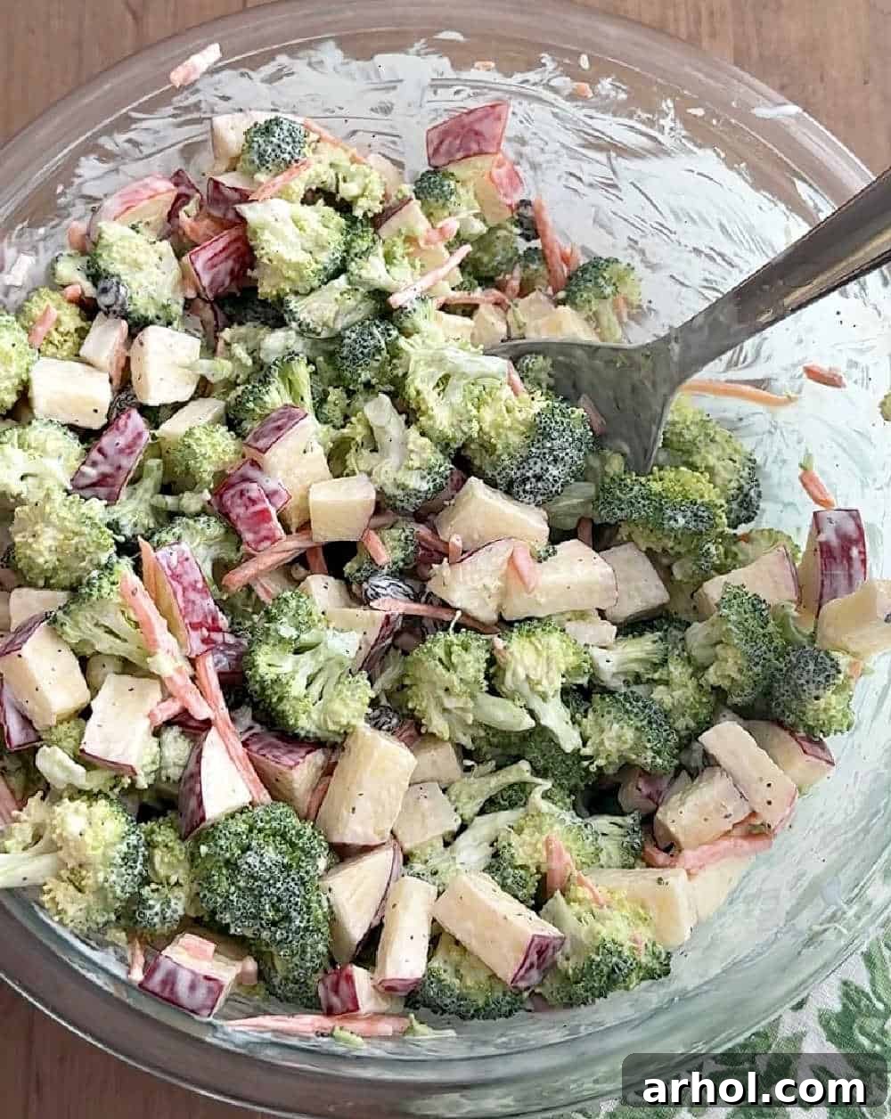 All the salad ingredients – broccoli, apples, carrots, and raisins – being mixed thoroughly with the creamy dressing in a large bowl.