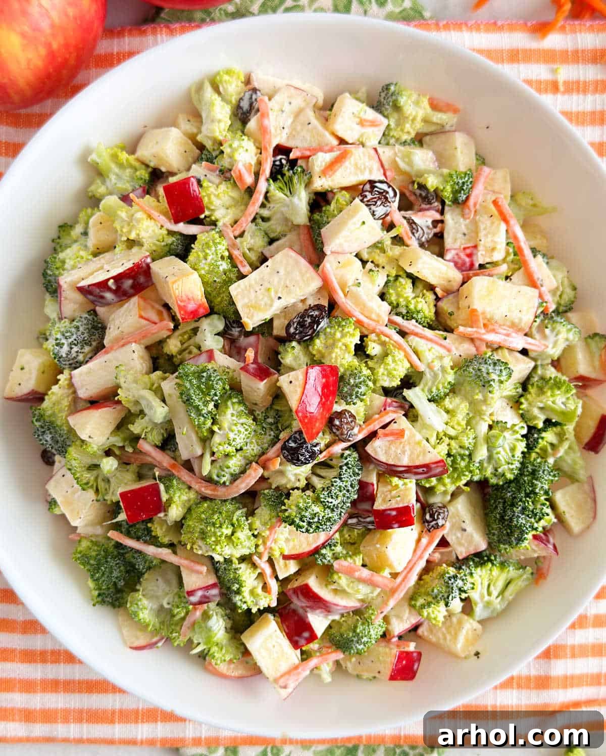 A close-up shot of a white bowl filled with freshly prepared broccoli apple salad, showcasing the texture and vibrant colors of broccoli, apples, carrots, and raisins.