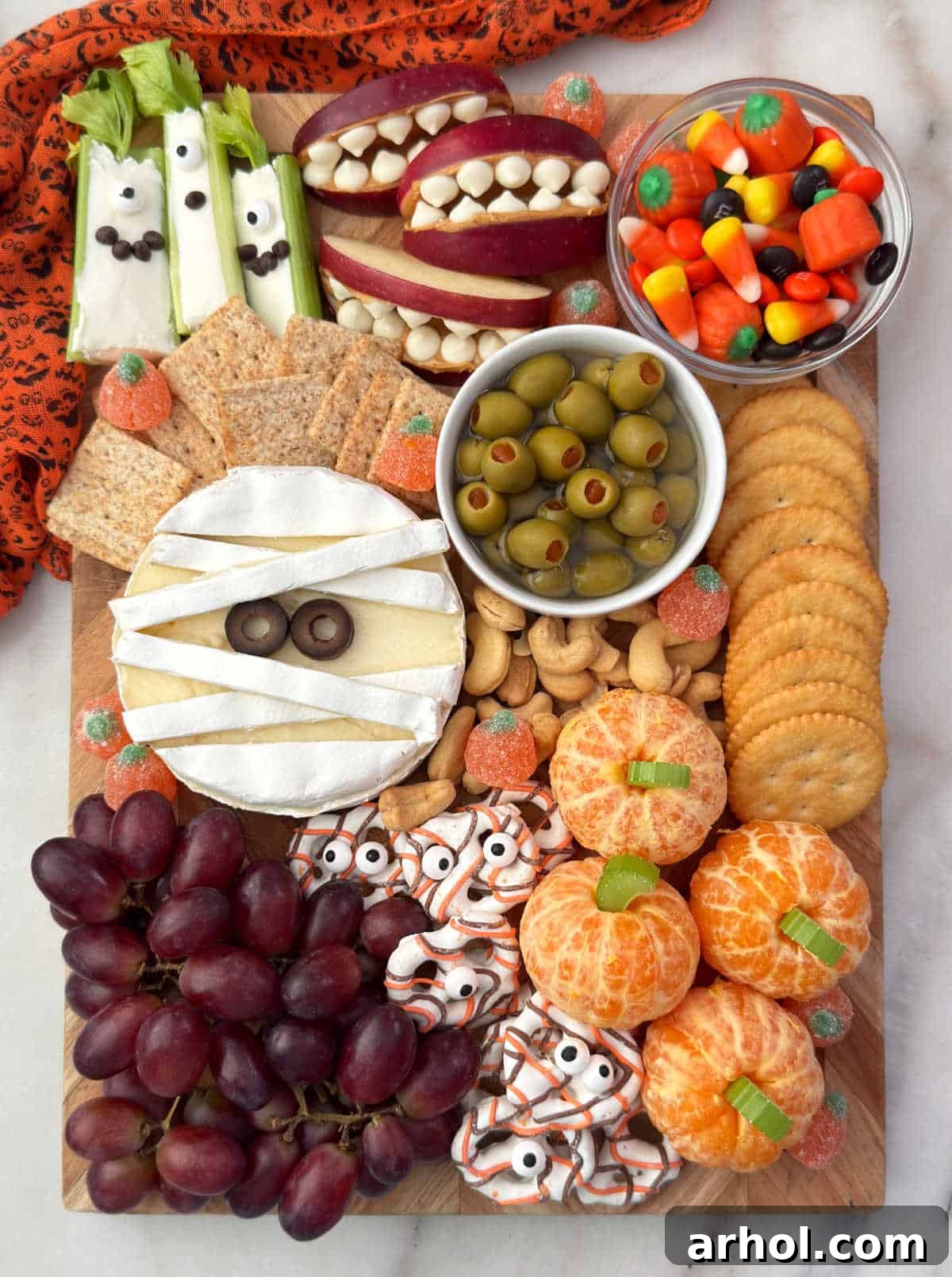 halloween charcuterie board with apple mouths, monster celery, brie mummy, pretzels, orange pumpkins, candy and olives.